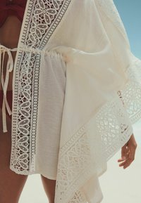 Cămașă albă cu detalii complicate din dantelă de-a lungul marginilor. Material ușor, talie strânsă cu șnururi, design fluid și margine cu model ondulat.