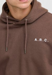 Haut à capuche marron avec cordon de serrage et embouts métalliques, portant le texte "A.R.C." en blanc sur le devant. Tissu doux avec un design décontracté.