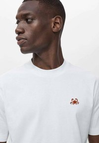 White cotton t-shirt with a ribbed crew neckline and a small embroidered crab design in red and orange positioned on the left chest.