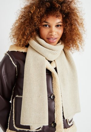 Jonge vrouw met krullend haar, gekleed in een bruine shearling jas en een lange beige gebreide sjaal, zacht glimlachend tegen een effen achtergrond.