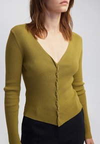 Cardigan lungo a maniche lunghe verde oliva a coste con scollatura a V profonda, caratterizzato da una fila di piccoli bottoni argentati e un orlo curvo.