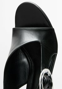 Zwarte leren sandalen met hoge hakken, voorzien van een brede band, open teen en een zilveren rond hardwaredetail. Glad textuur met uitsparingen aan de zijkanten.