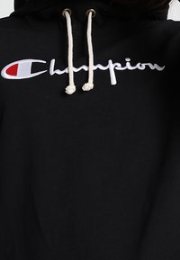 Zwarte hoodie van katoen, met een groot wit "Champion"-logo en een rood accent aan de linkerkant. Witte trekkoorden.