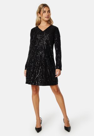 Abito nero a maniche lunghe con scollo a V, caratterizzato da un motivo testurizzato e decorazioni di paillettes. Modella che indossa tacchi a punta neri.
