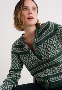 Rövid hajú nő zöld és fehér mintázatú, gombos kötött kardigánban, enyhén fölfelé nézve egy egyszerű háttér előtt.