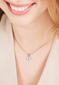 El collar de cadena plateada presenta un colgante en forma de ojo con acentos brillantes y una gema azul en el centro. Se lleva sobre una blusa beige.