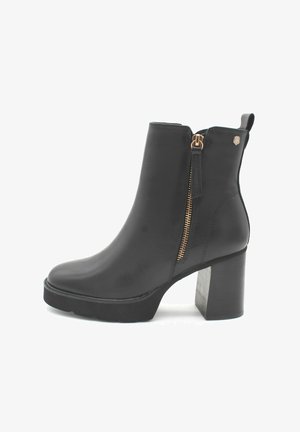Botín negro de cuero hasta el tobillo con tacón grueso, cremallera lateral con tirador de cuero y un pequeño logo metálico cerca del borde superior.