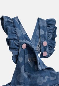 Salopette en denim avec motifs de cœurs, bretelles volantes et boutons roses. "GUES" brodé sur la poche avant. Couleur bleue avec des variations de texture.