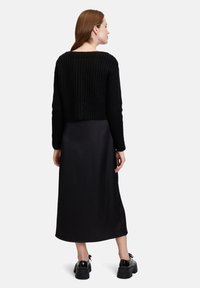Schwarzer, gestrickter, verkürzter Pullover kombiniert mit einem glatten schwarzen Midi-Rock. Das Model trägt klobige schwarze Schuhe und zeigt eine schlichte, aber moderne Silhouette.