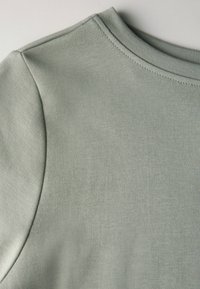 Helles grünes Oberteil aus Stoff mit gerippter Textur, rundem Ausschnitt und genähten Akzenten an den Schultern. Glattes und dehnbares Material.