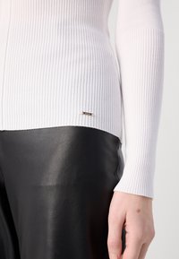 Gros plan d'une personne portant un haut blanc côtelé à manches longues et un pantalon en cuir noir, montrant une main avec une petite marque cutanée.