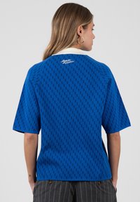 Mujer con cabello largo que lleva una blusa azul con estampado, mangas cortas, cuello blanco y pantalones grises con rayas finas, vista desde atrás.