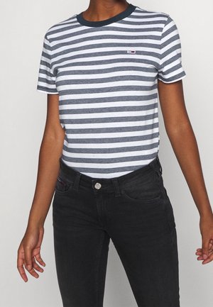 Personne portant une chemise à rayures bleu marine et blanche à manches courtes avec un jean noir sur fond uni.