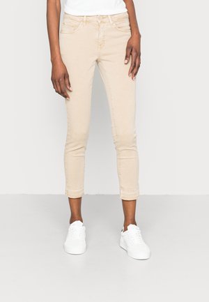 Beige cropped skinny farkut, joissa on etutaskut, hieno kulutusjälki ja vyölenkit, yhdistettynä valkoisiin lenkkareihin. Sileä rakenne, rento istuvuus.