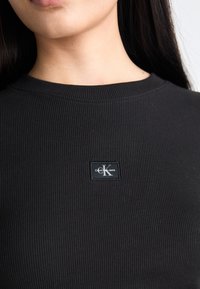 Black ribbed long-sleeve shirt featuring a rectangular logo patch with "Calvin Klein Jeans" and a prominent "K" in the center.