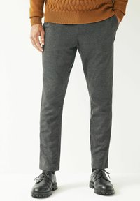 Mies, który nosi ciemnoszare slim-fit spodnie, czarne sznurowane buty i brązowy sweter, z dłońmi częściowo w kieszeniach, stoi na białym tle.
