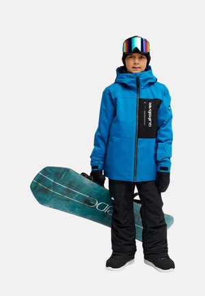 Enfant portant une veste de neige bleue, un pantalon de neige noir, des gants et des lunettes tenant un snowboard avec un motif turquoise.