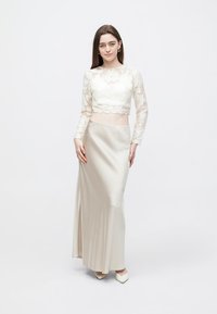 Λευκό crop top από δαντέλα με μακρύ μανίκι και κυματιστή λαιμόκοψη, σε συνδυασμό με μπεζ σατέν μακρύ φόρεμα με πλαϊνή σχισμή, φορεμένο με ανοιχτόχρωμες γόβες.