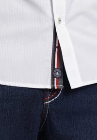 Weißes, geripptes Hemd mit einem geknöpften Kragen und einem Seitenetikett mit roten, weißen und blauen Akzenten, getragen über dunkelblauen Jeans mit kontrastierenden Nähten.