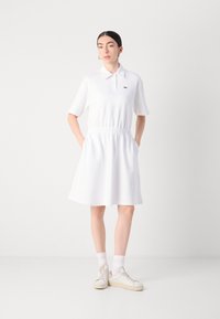 Balta polo suknelė su prigludusia juosmens linija ir trumpomis rankovėmis. Yra raukinėta apykaklė ir mažas logotipas krūtinėje. Lygus, kasdieniškas audinys.