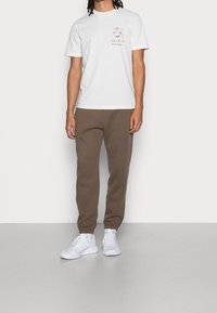 Persoon in een witte T-shirt met kleurrijke tekst, bruine joggerbroek en witte sneakers, staande tegen een effen lichte achtergrond.