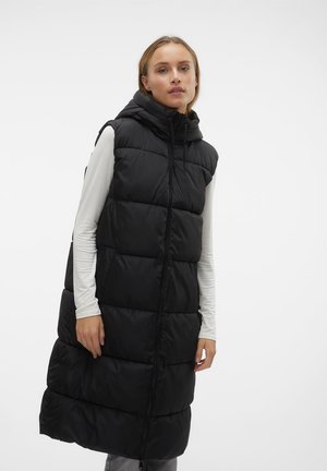 Femme portant une longue doudoune noire sans manches avec une capuche par-dessus une chemise blanche à manches longues, debout devant un fond blanc.
