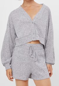 Woman wearing light gray knit two-piece loungewear set with long-sleeve cropped button-up top and drawstring shorts with pockets.