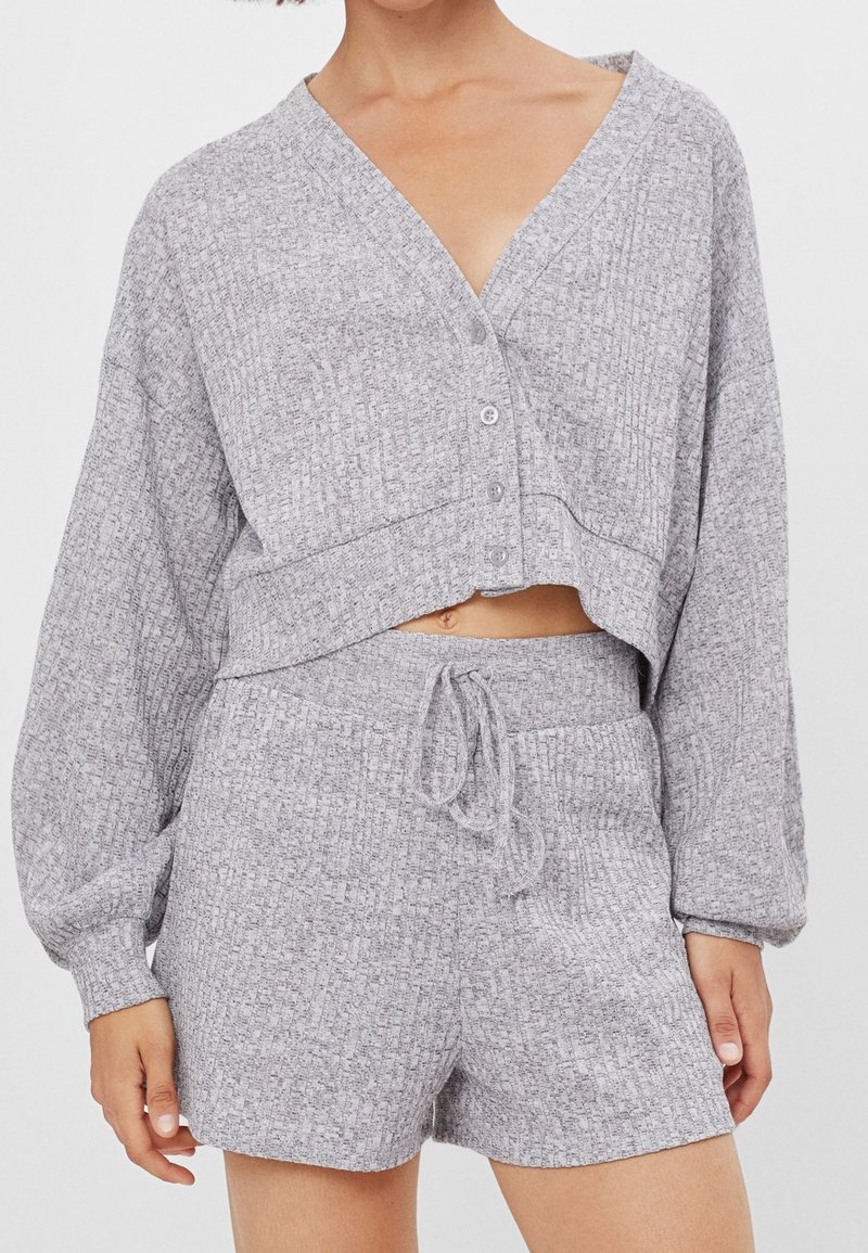 Woman wearing light gray knit two-piece loungewear set with long-sleeve cropped button-up top and drawstring shorts with pockets.