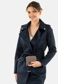 Cappotto trench in faux suede blu navy con vita cintata, colletto a revers e dettagli in metallo argentato, abbinato a un portafoglio con zip color taupe in mano.