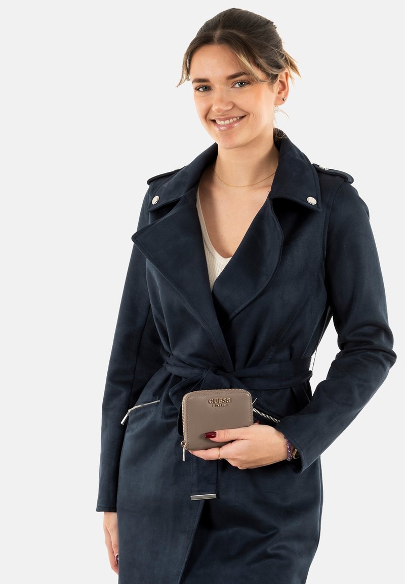 Cappotto trench in faux suede blu navy con vita cintata, colletto a revers e dettagli in metallo argentato, abbinato a un portafoglio con zip color taupe in mano.