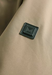 Beżowy materiał o gładkiej fakturze z kwadratową czarną skórzaną łatą i srebrnymi ćwiekami, z geometrycznym wzorem na środku.