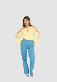 Blusa giallo chiaro con design a spalle scoperte, scollo a goccia e maniche ampie; abbinata a pantaloni aderenti blu e sandali bianchi.