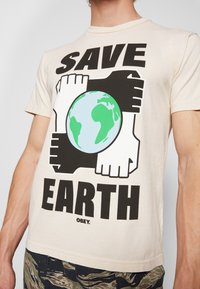 Béžové tričko s grafikou čtyř propletených černobílých rukou kolem zelené a modré Země a textem "ZACHRAŇME ZEMI."