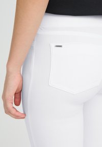 Balti šauri džinsi ar gludu tekstūru, ar aizmugurējo kabatu un nelielu logotipa birku. Vidukļa daļa ir vienkārša, bez redzamas aparatūras.