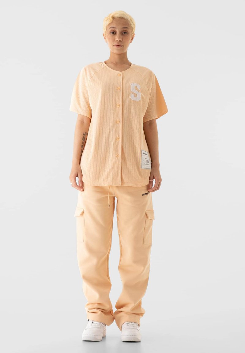 Viljalihvitud võrgumustriga ja valge tähega "S" virsikuvärvi lühikeste varrukatega nööpidega särk. Kanda sobivate virsikuvärvi taskupükste ja valgete tossudega.