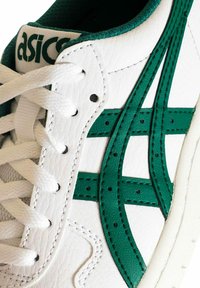 Zapatilla blanca de cuero con detalles en verde, con un acabado texturizado, rayas superpuestas y cordones blancos. Logo prominente en la lengüeta.