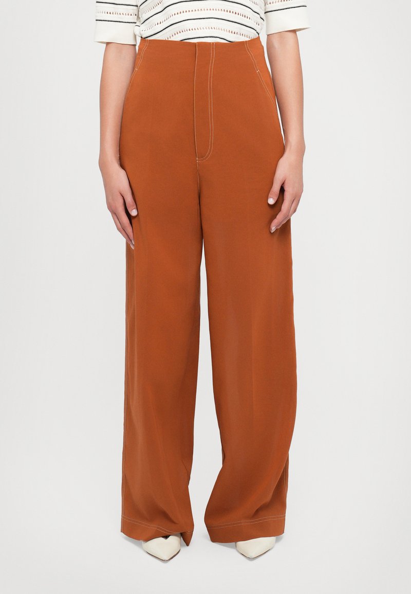 Weite Beinhosen aus rostorangefarbenem Stoff mit sichtbaren Nähdetails, kombiniert mit weißen Absatzschuhen. Glatte Textur, hoch geschnittenes Design.