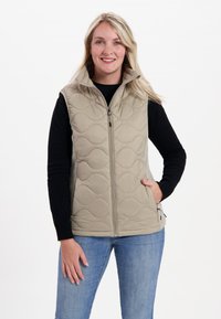 Beige gesteppte Weste mit hohem Kragen und Front-Reißverschluss; verfügt über Seitentaschen und eine weiche Textur, getragen über einem schwarzen Langarmoberteil.