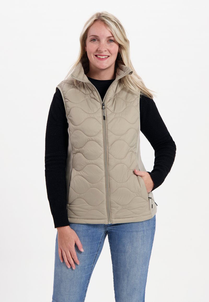Beige gesteppte Weste mit hohem Kragen und Front-Reißverschluss; verfügt über Seitentaschen und eine weiche Textur, getragen über einem schwarzen Langarmoberteil.