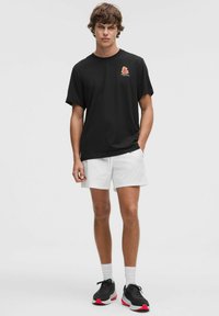 Tricou negru din bumbac cu un design floral pe partea stângă a pieptului, asortat cu pantaloni scurți albi și adidași negri cu accente roșii.