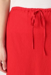 Raudona audinio sijonas su tekstūruota paviršiumi, turintis surinktą liemenį ir rišamą užsegimą. Dizainas apima paprastą, nepuošnų stilių.