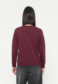 Persona con cabello oscuro y liso, vistiendo un sudadero burdeos de manga larga y jeans negros desgastados, visto desde atrás contra un fondo blanco.