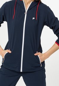 Mörkblå zip-up jacka med huva, vit dragkedja och F-logga, har röda förstärkningsdragskor och muddar, tillverkad av mjukt, lättviktigt tyg.