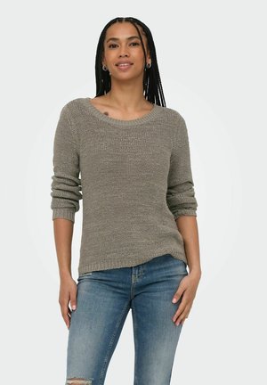 Femme aux cheveux tressés portant un pull en maille gris avec les manches retroussées et un jean bleu, debout, une main dans la poche, sur fond blanc.