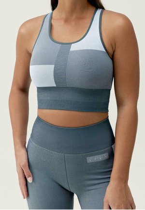 Vrouw draagt een blauw-grijze naadloze sportbeha en hooggetailleerde legging, staande met één hand op haar dij rustend.