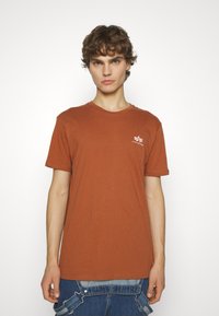 Kupferfarbiges Baumwoll-T-Shirt mit Rundhalsausschnitt, kurzen Ärmeln und einem kleinen weißen Logo auf der linken Brust. Glatte Textur, lockere Passform.