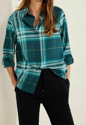 Femme portant une chemise à carreaux vert sarcelle et bleu avec les manches retroussées et un pantalon noir, les mains dans les poches.