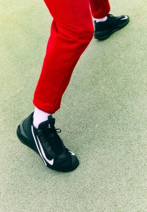 Personne portant un pantalon rouge et des chaussettes blanches marche sur un sol vert texturé avec des chaussures de sport Nike noires.