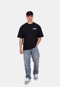 T-shirt noir surdimensionné avec le logo "CRYSTAL", porté avec un jean bleu clair et des baskets grises. Texture plate, manches courtes et coupe ample.