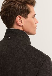 Czarny wełniany płaszcz w kolorze ciemnoszarym z wysokim kołnierzem, wykonany z teksturowanej tkaniny i z małą metką z logo blisko kołnierza. Dopasowany krój i czyste linie.